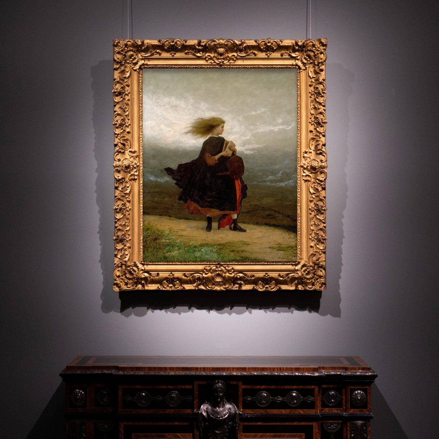 A fine art print in a very ornate gold frame hangs on a dark coloured wall. The picture depicts a windswept young lady walking across a landscape in a velvet coat and holding a book.