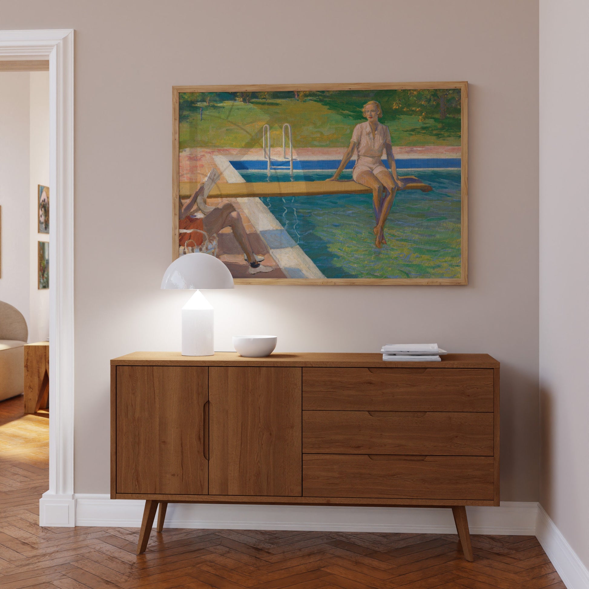 A fine art print of a beautiful Art Deco portrait in oil of a young lady resting on a spring board above a swimming pool. The print is displayed hanging on the wall in a light-wood picture frame in a hallway above a side table.