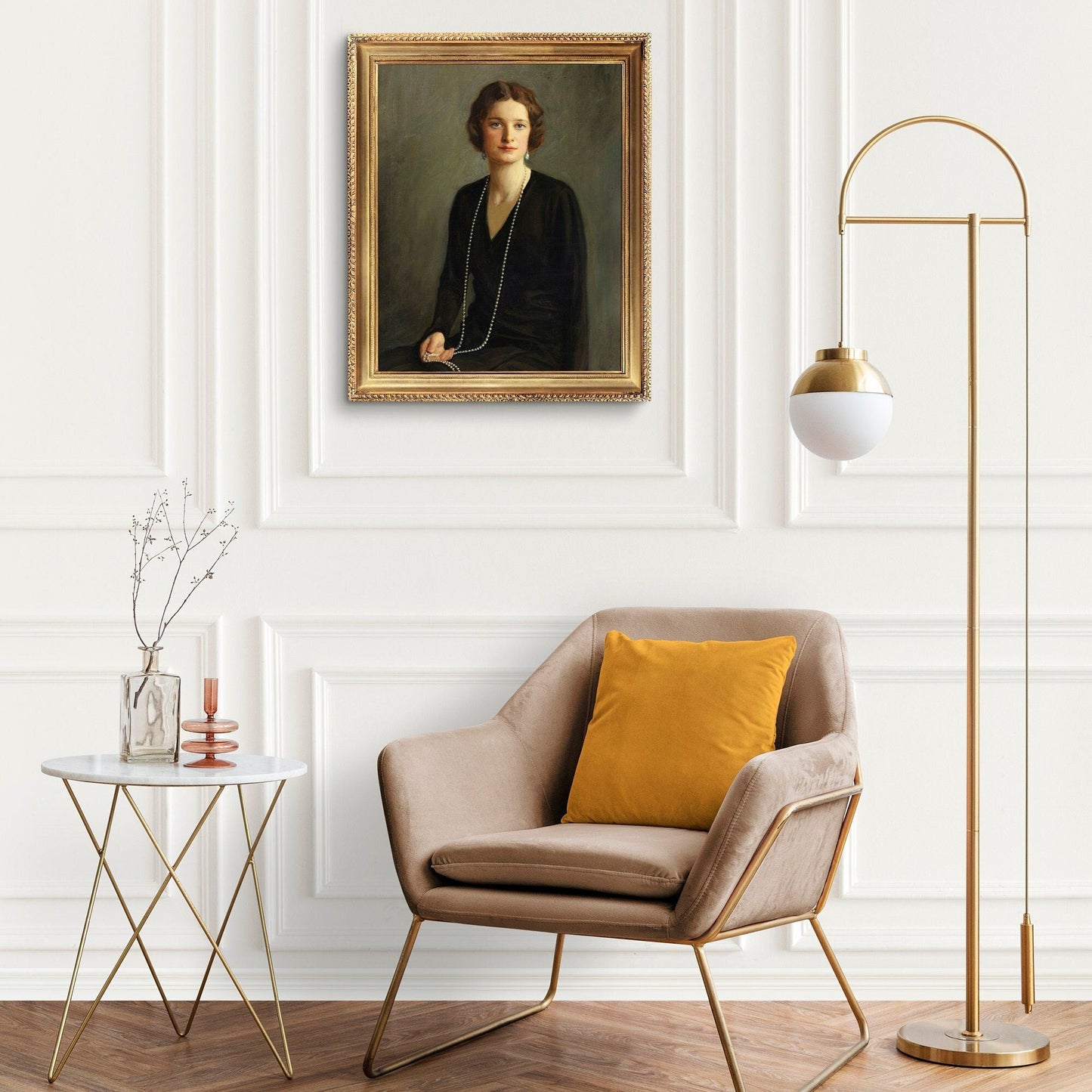 A fine art print of a beautiful portrait from the Art Deco period. The print is displayed hanging on a white panelled wall in a gold picture frame, next to an Art Deco lamp and chair with gold legs and a yellow cushion.