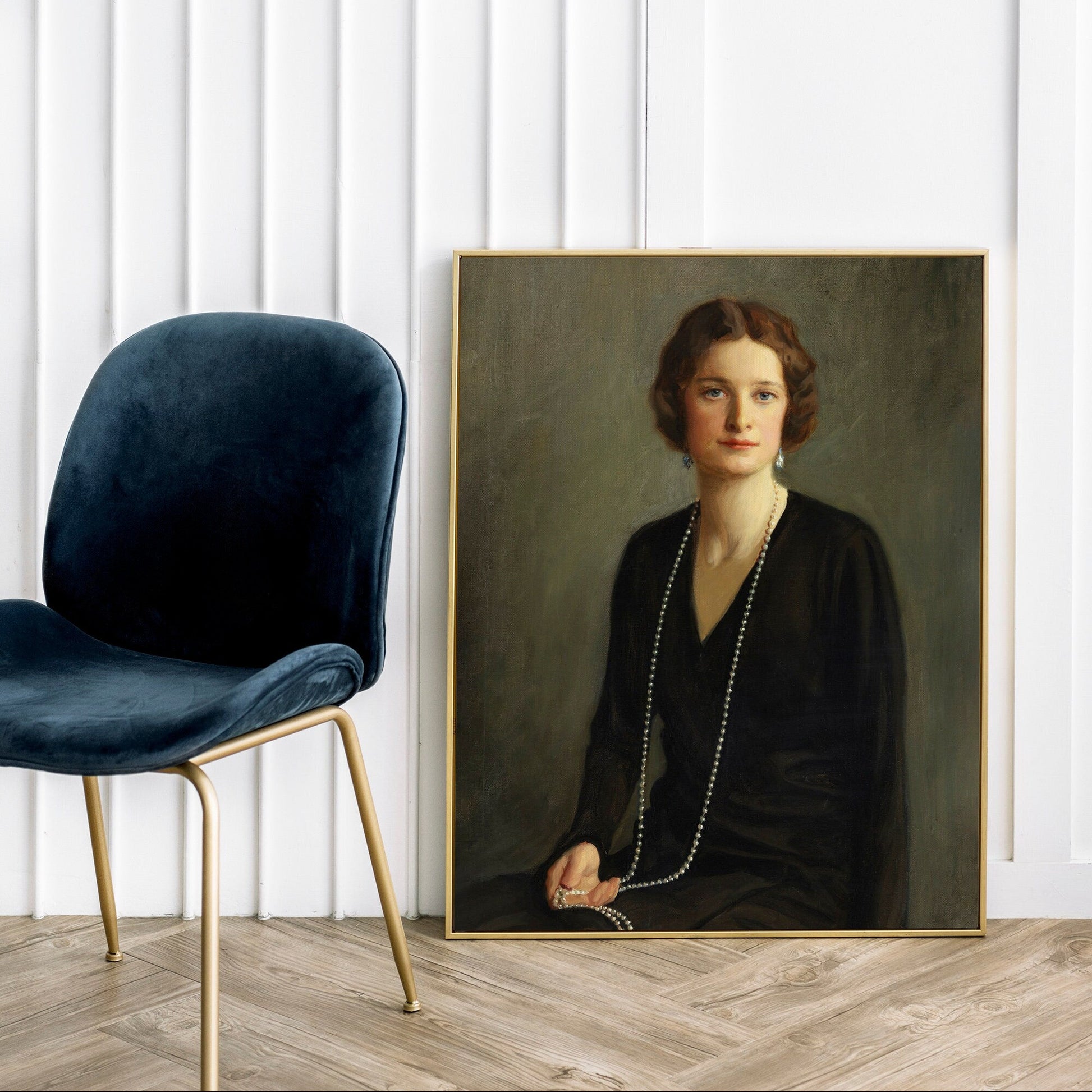 A fine art print of a beautiful portrait from the Art Deco period. The print is displayed resting against a white panelled wall in a thin gold picture frame, next to a blue velvet chair with gold legs.