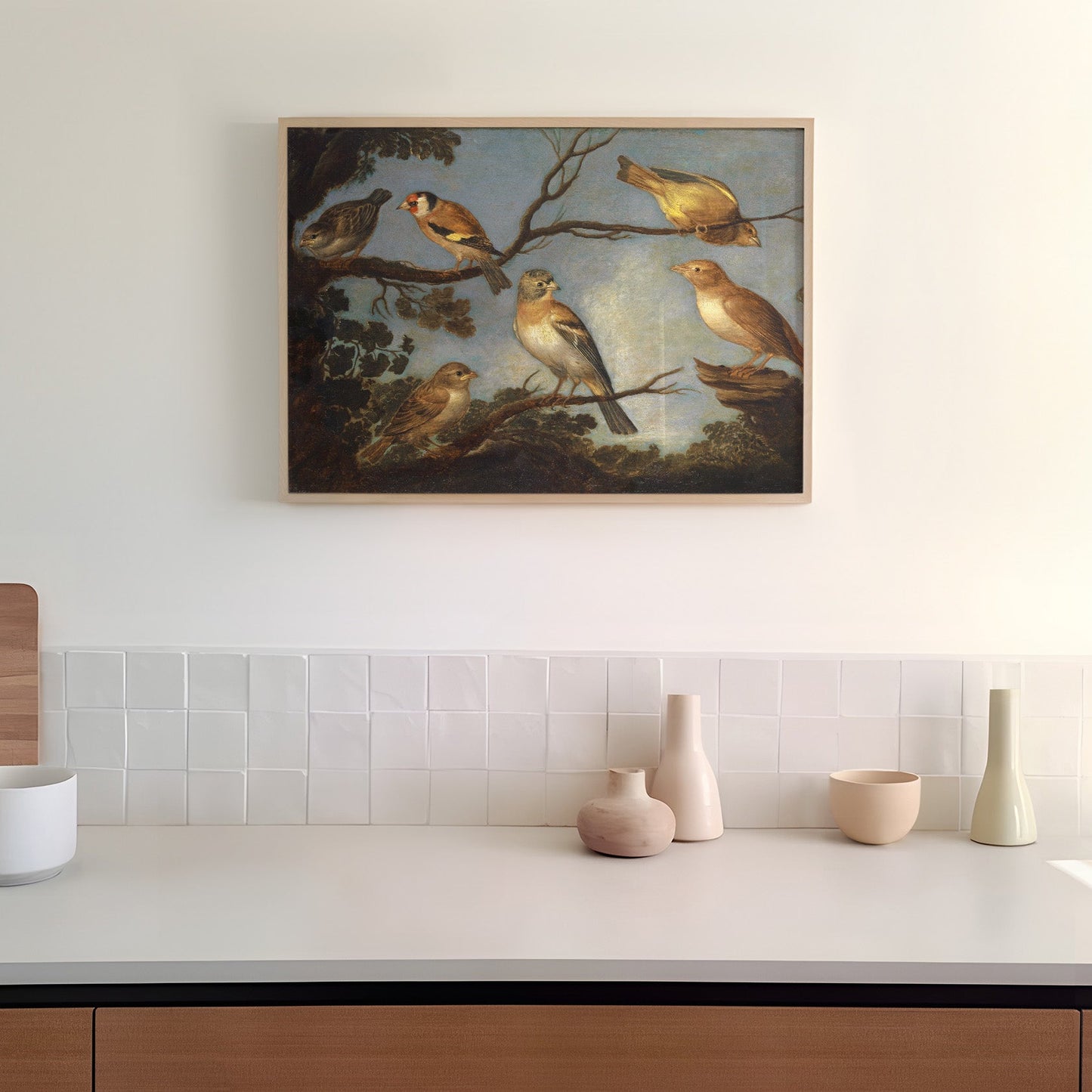 A framed print of garden birds gathered together on the branches of a tree. The print is displayed hanging on the wall in a kitchen in a wooden picture frame.