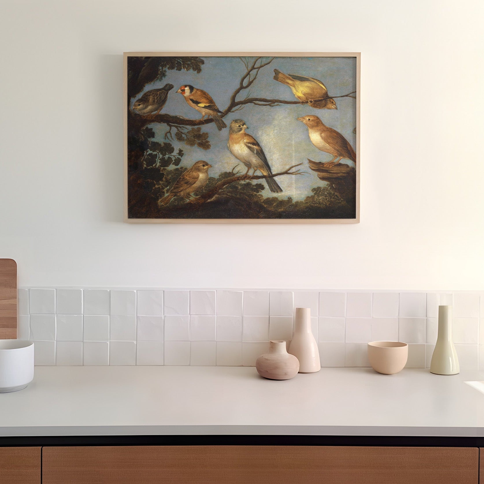A framed print of garden birds gathered together on the branches of a tree. The print is displayed hanging on the wall in a kitchen in a wooden picture frame.