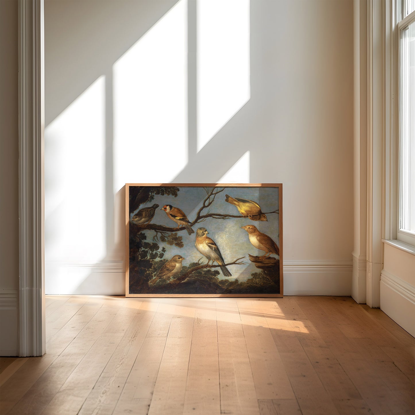 A framed print of garden birds gathered together on the branches of a tree. The print is displayed resting against a wall in a wooden picture frame with light streaming through the window.