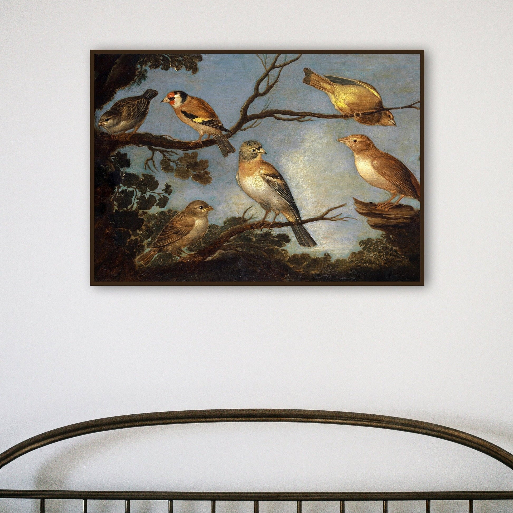 A framed print of garden birds gathered together on the branches of a tree. The print is displayed hanging on the wall in a thin black picture frame above a metal headboard.