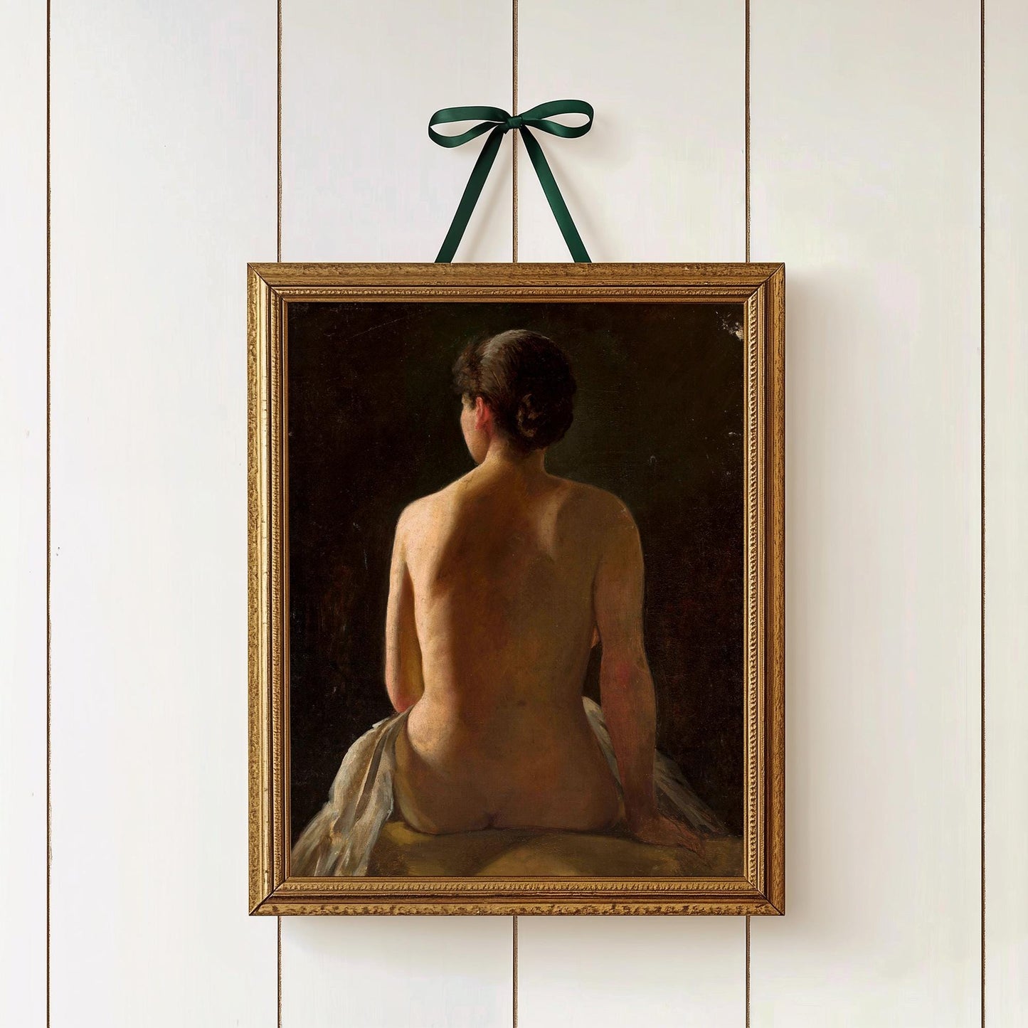 A fine art print of a painting of a seated female nude, shown from behind. The print is displayed in a wooden picture frame, hung by a green ribbon in a bow.