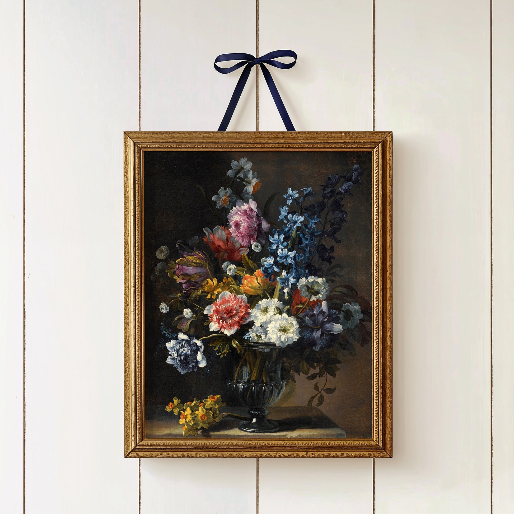 Gold-framed print of a floral arrangement on a white paneled wall, hung with a ribbon.
