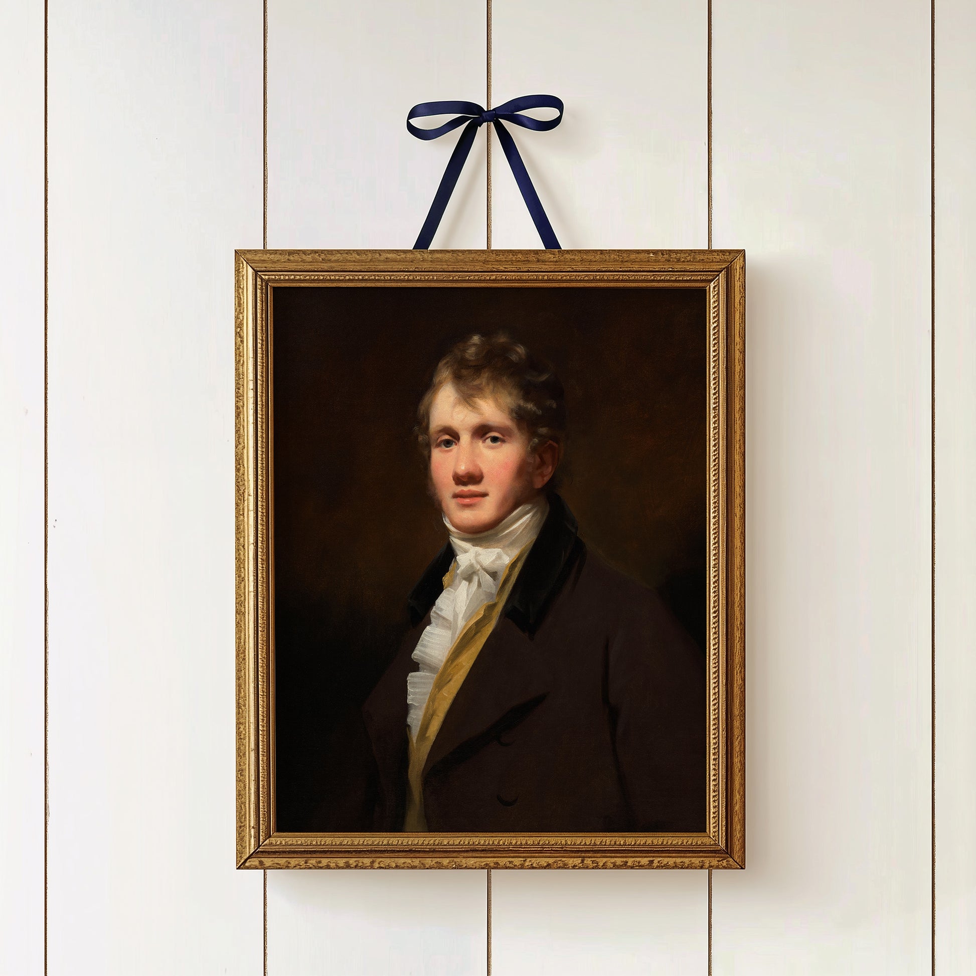 Framed portrait of a man hanging on a white paneled wall in a gold frame with a ribbon.