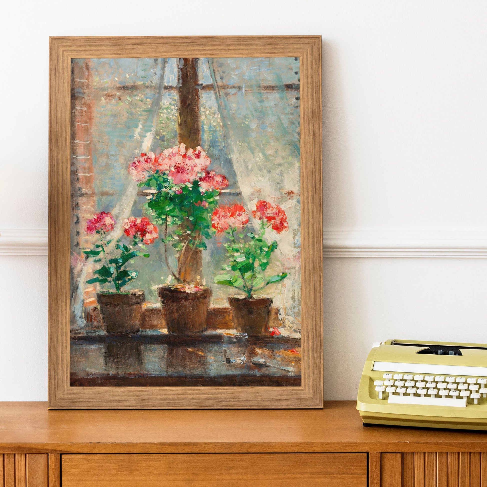 A picture of red and pink geraniums in terracotta pots sat on a windowsill, is framed in a light wood picture frame, and sits on top of a cabinet next to a typewriter.