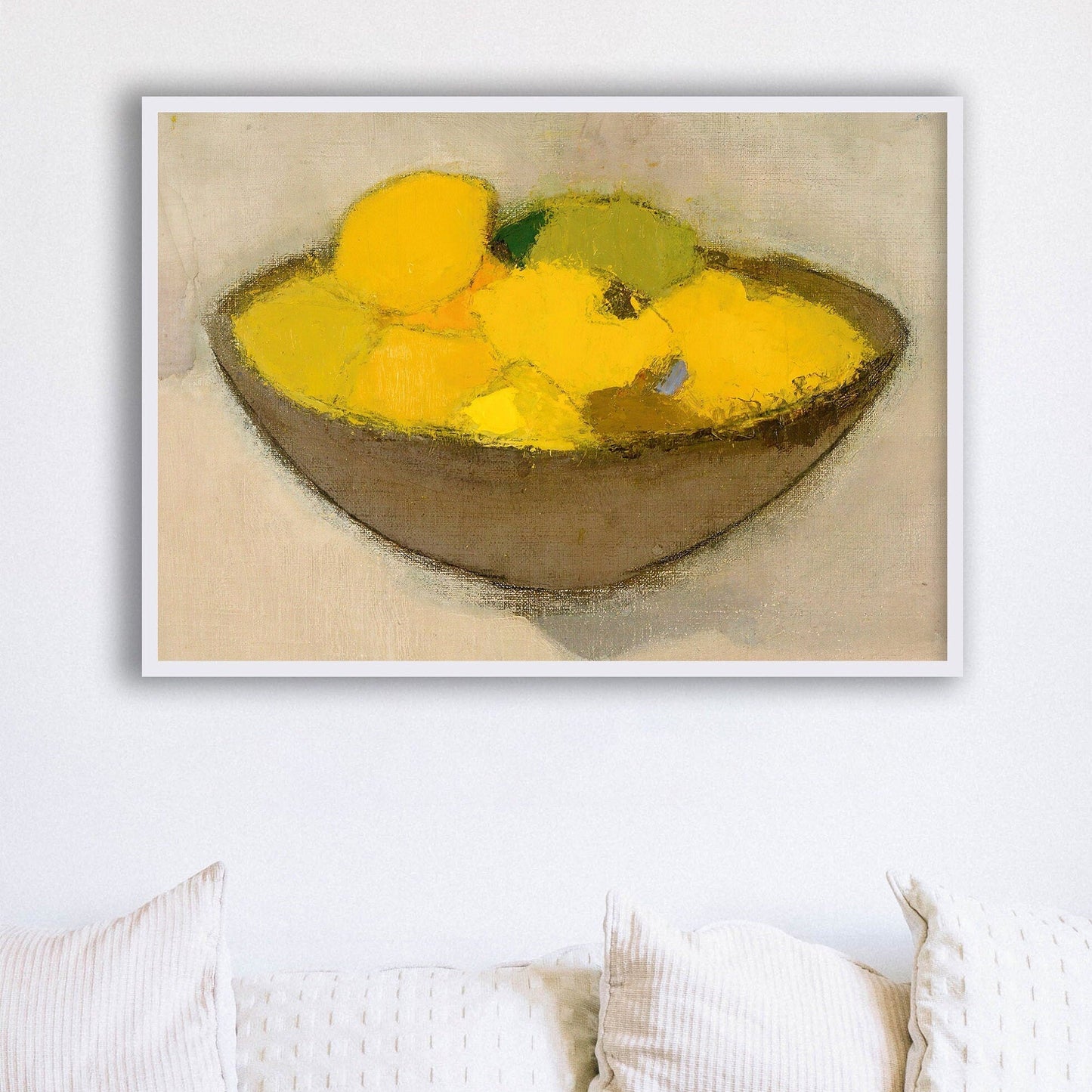 Framed print of a bowl with yellow lemons on a neutral background, hung above some white cushions.