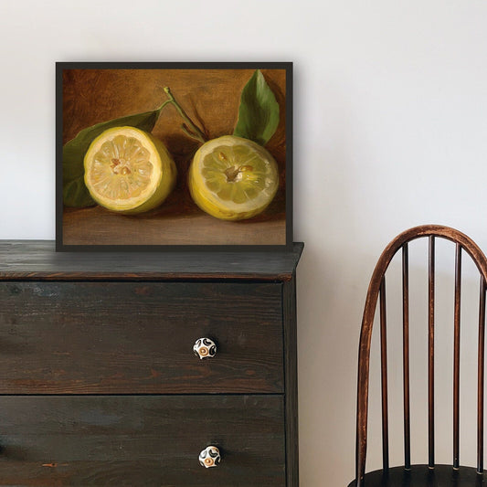 A fine art print of a beautiful 19th Century oil painting of a simple still life of a halved lemon with its leaves. The print is displayed resting on top of a dark chest of drawers in a thin black picture frame.
