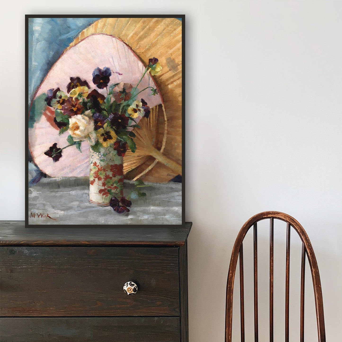 Wooden chair in front of a black dresser with a framed fine art print of a floral still-life on top.