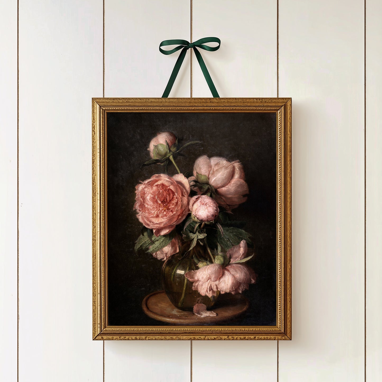 A gold framed fine art print of pink peonies on a dark background, hung on a white wall with a green ribbon