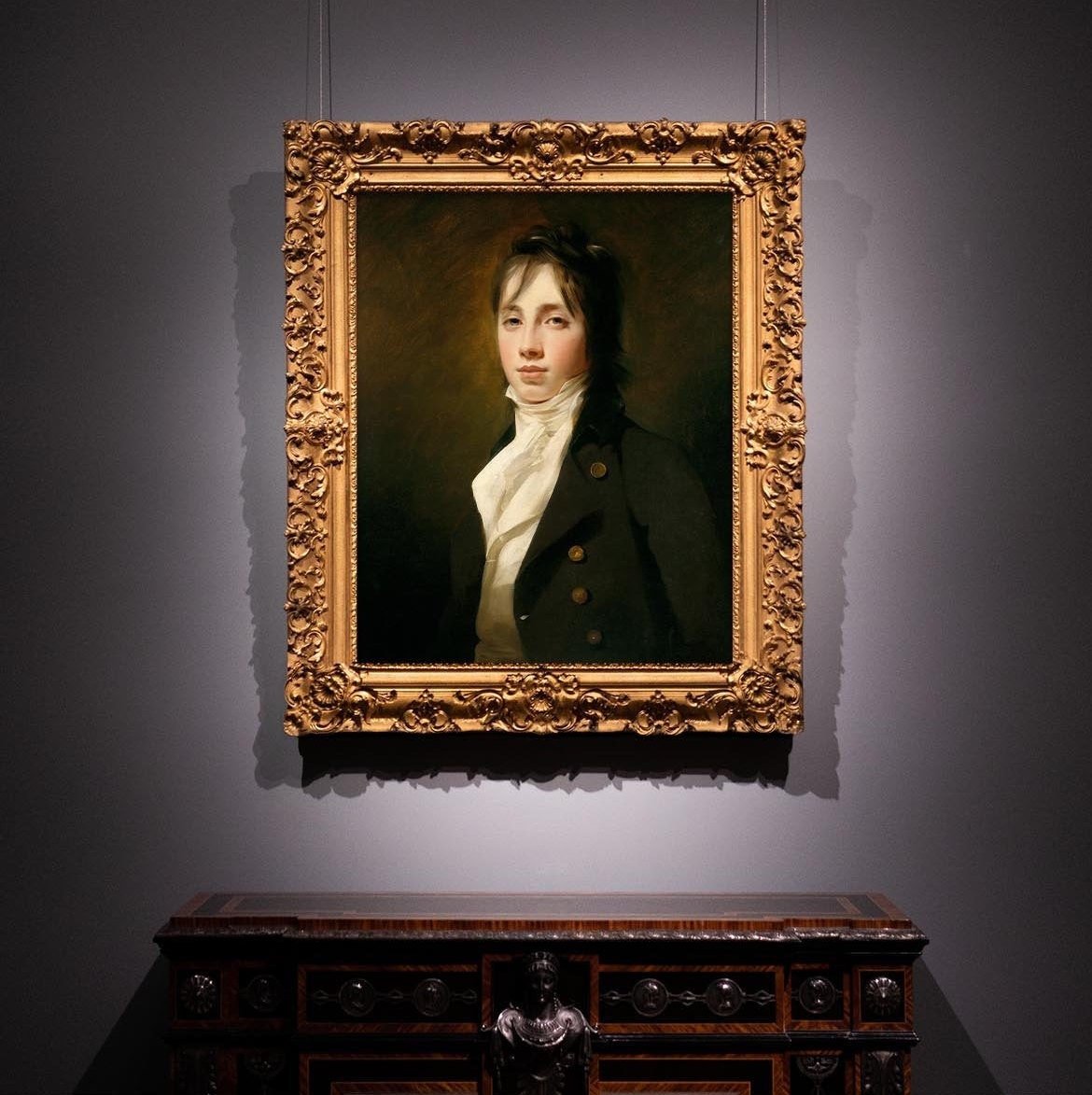 Portrait of a man in an ornate gold frame on a gray wall with a wooden cabinet below.